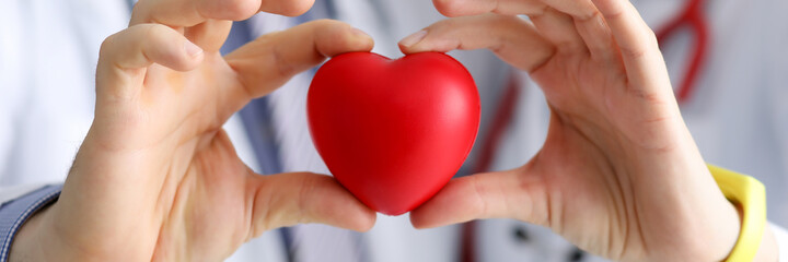 Doctors cardiologists hold red toy heart in clinic closeup. Diagnosis and treatment of myocardial infarction concept.