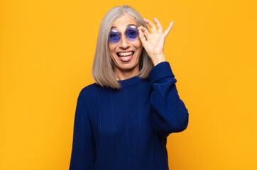middle age woman smiling happily with funny face, joking and looking through peephole, spying on...