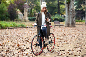 Beautiful young woman sending messages with her smart phone while cycling through the park in autumn.