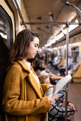 Fototapeta na wymiar young woman in autumn coat reading newspaper while traveling in metro train