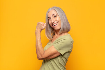 middle age woman feeling happy, satisfied and powerful, flexing fit and muscular biceps, looking...