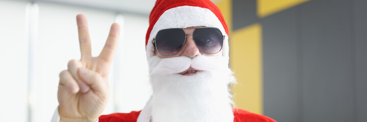 Man in santa claus costume with white beard and sunglasses holds his fingers in the sign of...