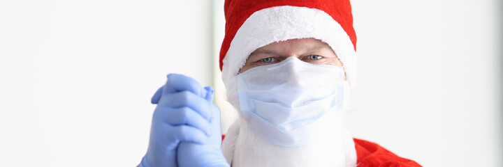 Man in santa claus costume and protective medical mask and gloves stands with his hands together and smiles. Safe New Year and Christmas celebration in coronavirus pandemic concept