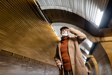 Fototapeta na wymiar elderly man in autumn clothes touching eyeglasses while standing on metro platform
