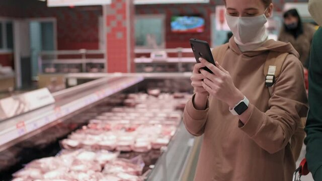 Medium slow-mo close-up of girl in protective mask taking picture of food products on smartphone showing photo to her male friend