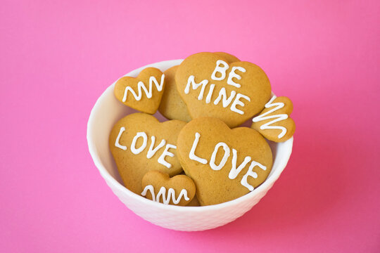 Plate with homemade freshly baked cookies in heart shape with icing and words of Love and Be Mine on pastel pink background. Valentines Day treat, sweet gift, romantic bakery. Angagement and wedding.