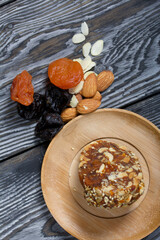 An energy bar made of dried fruits, nuts and seeds. Nearby there are ingredients for cooking, dried apricots, almonds and prunes. On black pine boards.