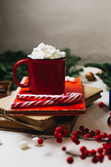 A red mug with cocoa and marshmallows stands on a stack of old books. Nearby are spruce branches, New Year's decorations, a branch of ikles and stamps for letters. Christmas mood and a hot drink.