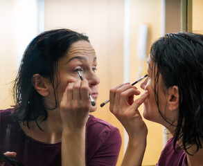 A middle-aged woman are doing morning makeup in front of a large mirror.