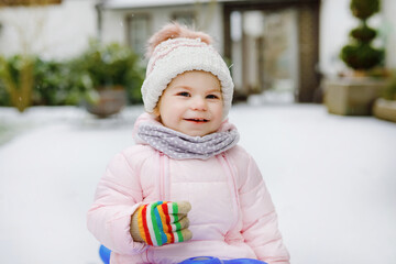 Cute little toddler girl enjoying a sleigh ride on snow. Child sledding. Baby kid riding a sledge in colorful fashion clothes. Outdoor active fun for family winter vacation on day with snowfall