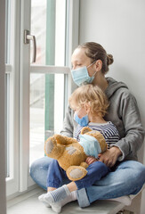 Mother and child in home quarantine looking out the window, wearing a medical mask against viruses during coronavirus COVID-2019 and flu lock down. Selective focus