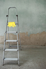 Old folding gray metal ladder and yellow rubber gloves on the background of a dirty construction site. Household repair, construction. Selective focus, artificial noise. Color of the year 2021 © Veaceslav