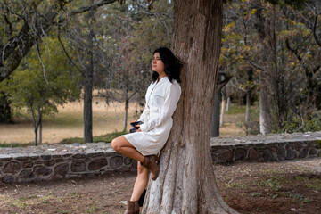 Mujer joven con vestido blanco apoyada en un árbol revisando su teléfono celular