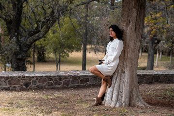 Mujer joven con vestido blanco apoyada en un árbol revisando su teléfono celular