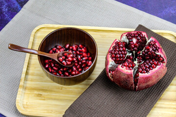 Wooden bowl containing wooden spoon and red pomegranate seeds. Pomegranate fruit open divided into five parts held together on a napkin / cloth on wooden tray on a blue background. Fruit pomegranate.