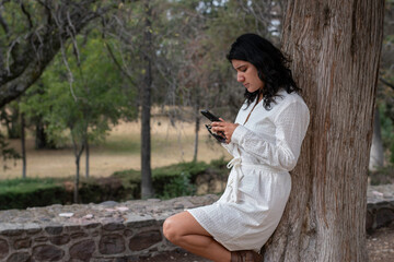 Mujer joven con vestido blanco apoyada en un árbol revisando su teléfono celular
