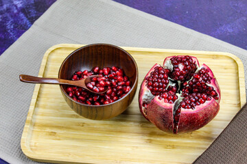 Wooden bowl containing wooden spoon and red pomegranate seeds. Pomegranate fruit open divided into five parts held together on a napkin / cloth on wooden tray on a blue background. Fruit pomegranate.