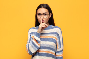 young hispanic woman asking for silence and quiet, gesturing with finger in front of mouth, saying...