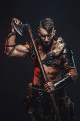 Fototapeta premium Barbaric and grimy northern vandal in light armour with fur holding his huge axe on his shoulder in dark background.