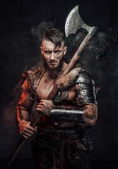 Fototapeta na wymiar Muscular and awesome viking fighter armed with huge axe and dressed in light armour poses in smokey dark background.