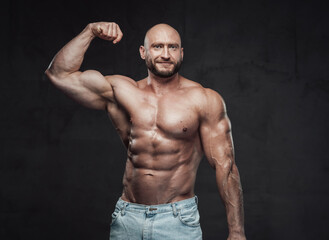 Obraz na płótnie Canvas Portrait of a happy and muscular guy with bald head and naked torso posing in dark background showing his huge biceps looking at camera.