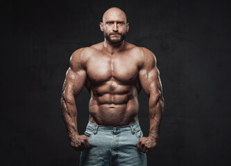 Obraz na płótnie Canvas Muscular and bald caucasian man with perfect abs and huge biceps poses in dark background looking at camera with serious face.