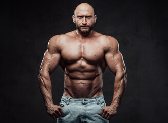 Fototapeta na wymiar Bearded and hairless man with powerful and muscular build in jeans shorts poses with serious face in dark background looking at camera.