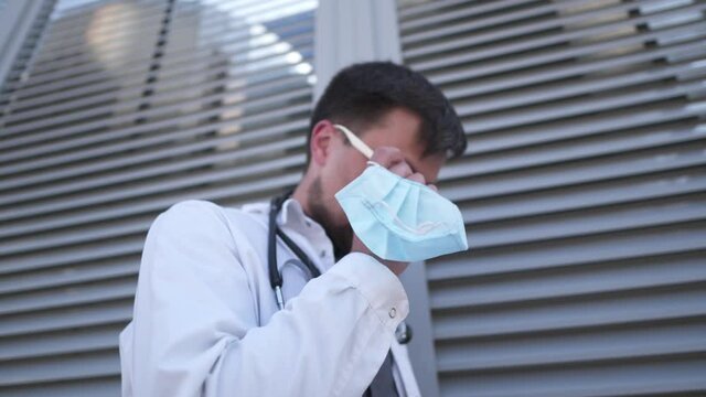 Pandemic coronavirus and covid 19. Serious young Caucasian doctor puts on mask again before returning to see patients at clinic. Physician sadness fatigue stress and exhaustion after major operation