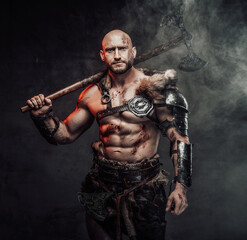 Fototapeta na wymiar Violent scandinavian barbarian with muscular nude build and bald head posing in dark atmospheric background with smoke with his huge two handed axe.
