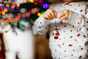 Adorable toddler girl in pajamas decorating Christmas tree with toy in cute hands. Little child in nightwear standing by Xmas tree. celebration of traditional family winter holiday