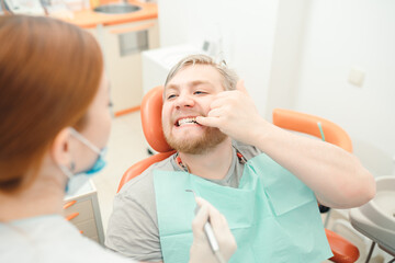 A man sits in the dentist's chair and points his finger at the doctor with a sore tooth. Dentistry concept, teeth and gum health