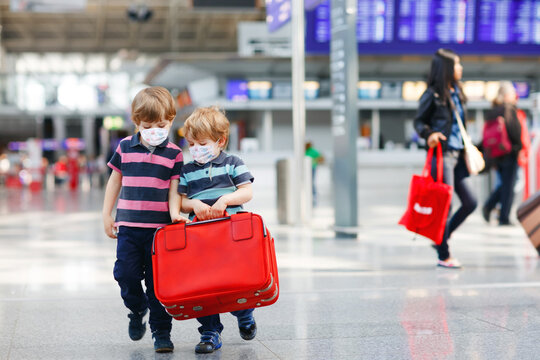 Two little sibling boys wear medical mask and going on vacations trip with suitcase at airport, indoors. Happy twins brothers walking to check-in or boarding for flight during corona virus pandemic