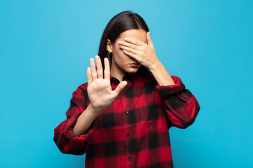 young hispanic woman covering face with hand and putting other hand up front to stop camera,...