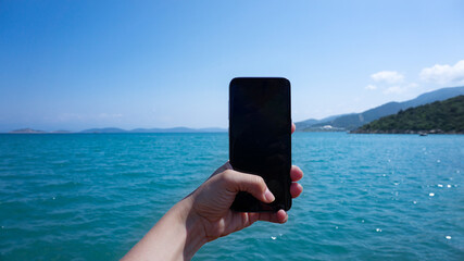 Vertical smartphone. Tourist hand holding a black blank space screen smart phone while in vacation in the beach with view of the water blue landscape. Coast of Turkey, Aegean Sea
