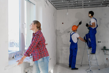 A woman stands by the window and examines the diagram of the house. Two builders drill a wall while...