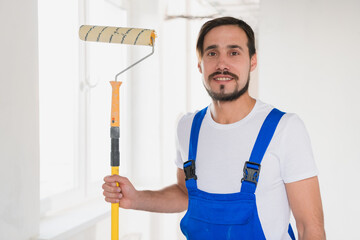 A bearded repairer in a blue overalls holds a roller in his hand and poses at the camera