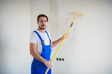 A handyman in a blue overalls holds a roller in his hands, smiles and looks at the camera