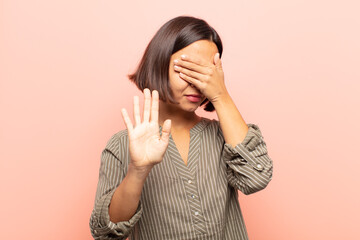 young hispanic woman covering face with hand and putting other hand up front to stop camera,...