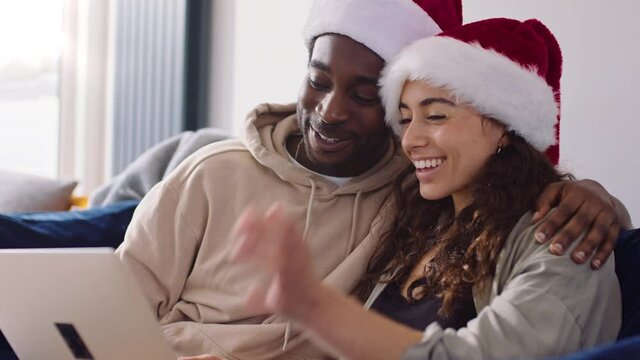 Young Couple Wearing Santa Hats At Home Making Video Call On Laptop Computer At Christmas