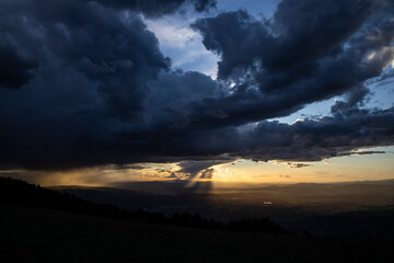 From the top of a mountain, there is a wide view during sunset time (golden hour). The gray clouds full of rain collide with the sunlight creating suggestive contrasts with plays of light.