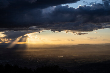 From the top of a mountain, there is a wide view during sunset time (golden hour). The gray clouds full of rain collide with the sunlight creating suggestive contrasts with plays of light.