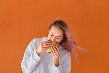 A young woman with pink hair eats delicious food. Big Burger. The concept of harmful nutrition. Orange background.