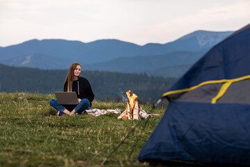 young female freelancer working on laptop in the mountains in the evening. Tourist girl sitting near campfire and tent. Copy space