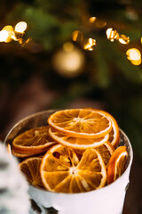 Vertical photo with a cup of dried orange slices on a dark background of a Christmas tree with bokeh from garlands.