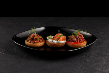 Three round bruschetta with different flavors and ingredients (fish, vegetables, meat) on dark background