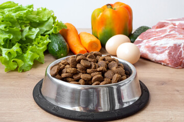 Dry pet dog food with natural ingredients. Raw meat, vegetables, eggs and salad near bowl with dry pet feed on wooden background. concept of pet correct balanced and healthy nutrition