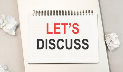 The word let's discuss is written in a notebook that sits on a gray desktop along with a laptop.