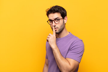 young handsome man asking for silence and quiet, gesturing with finger in front of mouth, saying...