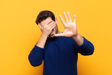 young handsome man covering face with hand and putting other hand up front to stop camera, refusing...