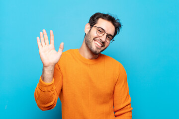 young handsome man smiling happily and cheerfully, waving hand, welcoming and greeting you, or...